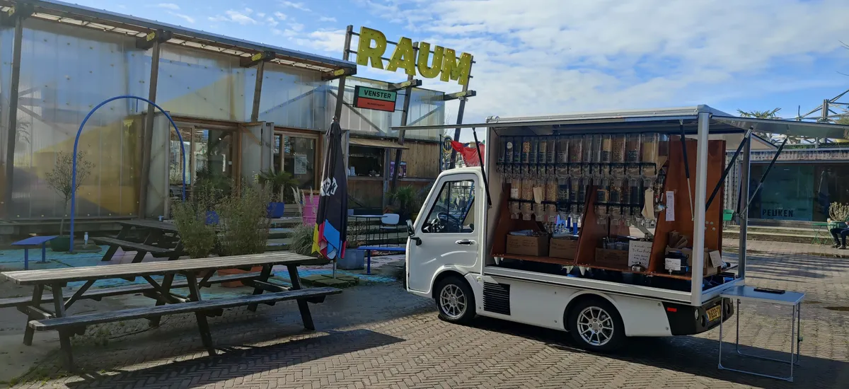 Een mobiele winkel van LOOS staat in een open binnenplaats. Het voertuig heeft verschillende dispensers en bakken voor producten. Klanten kunnen hun eigen bakjes vullen.